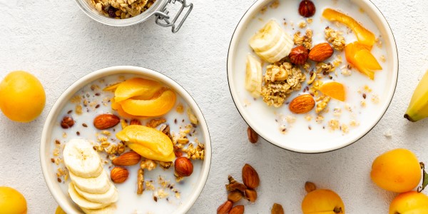 Two bowls of rice and oat cereal extracts are filled with dairy alternative milk and fresh fruit, displayed on a table alongside fruit and nuts.