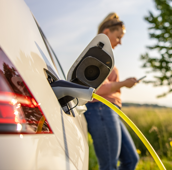 An electric car plugged in and charging outside with a woman on her phone waiting in the background.