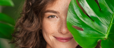 A woman covers the left side of her face with a large monstera plant leaf